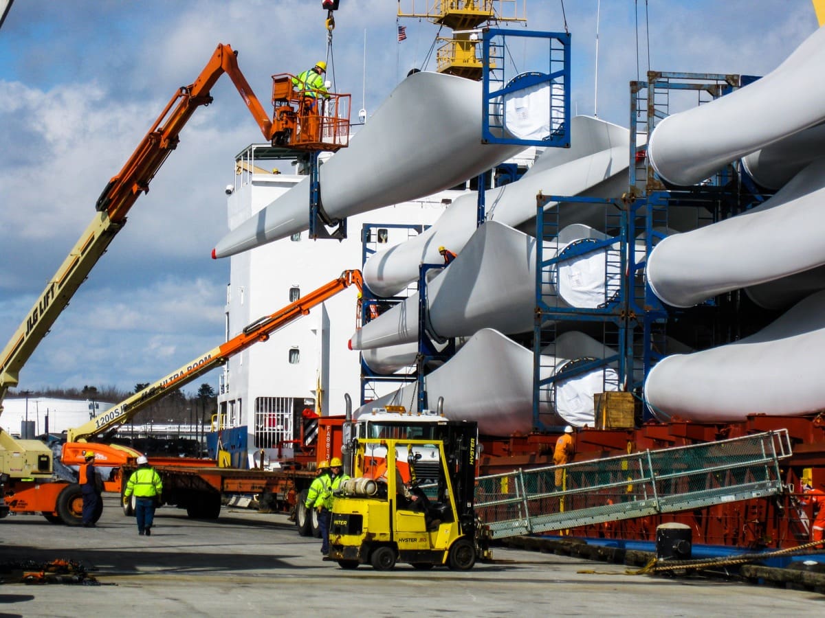 Workers use cranes and lifts to load large wind turbine blades onto a cargo ship at a port. The blades are secured in blue steel racks, and the team is wearing safety gear and high-visibility jackets. A forklift and multiple boom lifts are active on the dock under a partly cloudy sky.