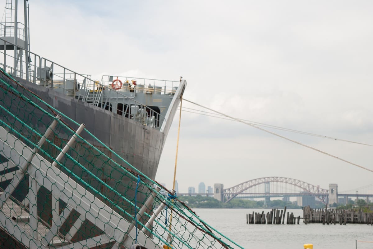 Close-up view of the bow of a large cargo ship docked at a pier, with gangways connected for boarding. The background features a calm river, distant city skyline, and an arched bridge under an overcast sky.