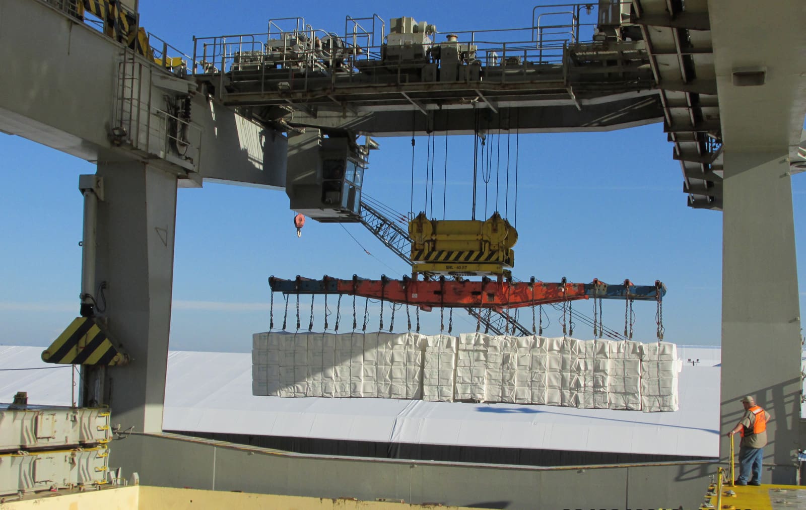 Heavy machinery and industrial equipment operating at a shipping terminal. A yellow loader and white dump truck are positioned beneath large steel hoppers, with cranes and conveyor systems visible in the background against a blue sky.