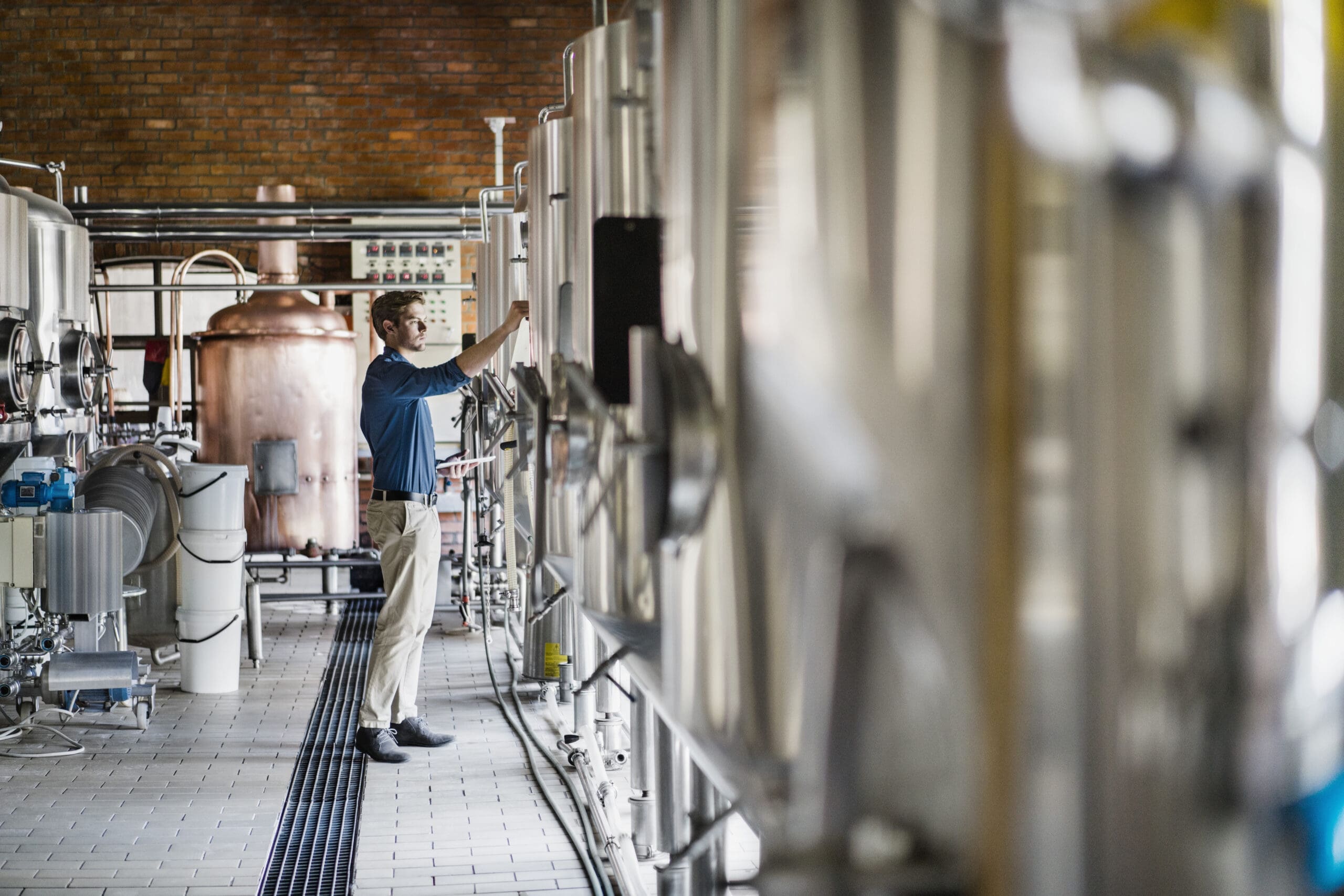 Male worker operating machinery in brewery. Full length of professional is holding digital tablet while working amidst metallic vats. He is in beer industry.