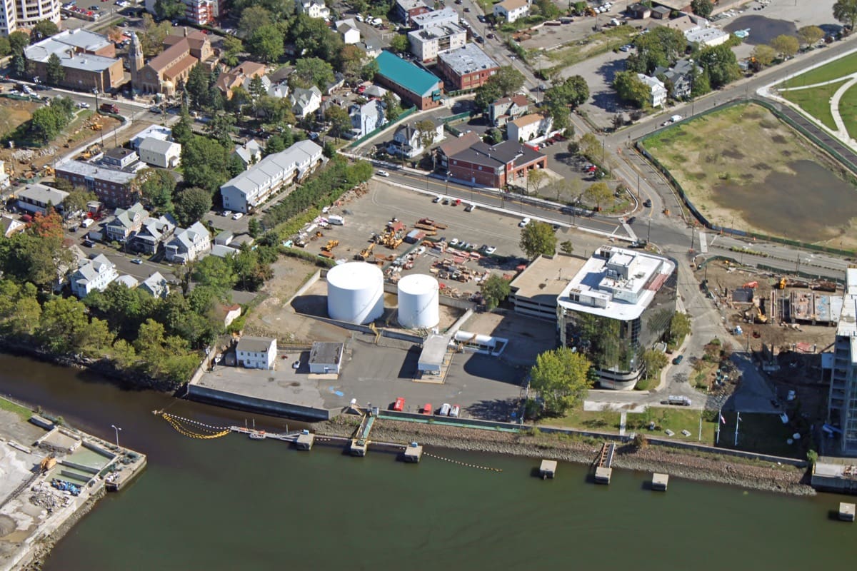 Stamford-Terminal_resized Aerial view of a small industrial site with two large white cylindrical storage tanks near the waterfront. The site is adjacent to a river or canal with a small dock and several concrete piers. Surrounding the site are commercial buildings, parking areas, and a residential neighborhood with houses and trees. Roads and green spaces are also visible.