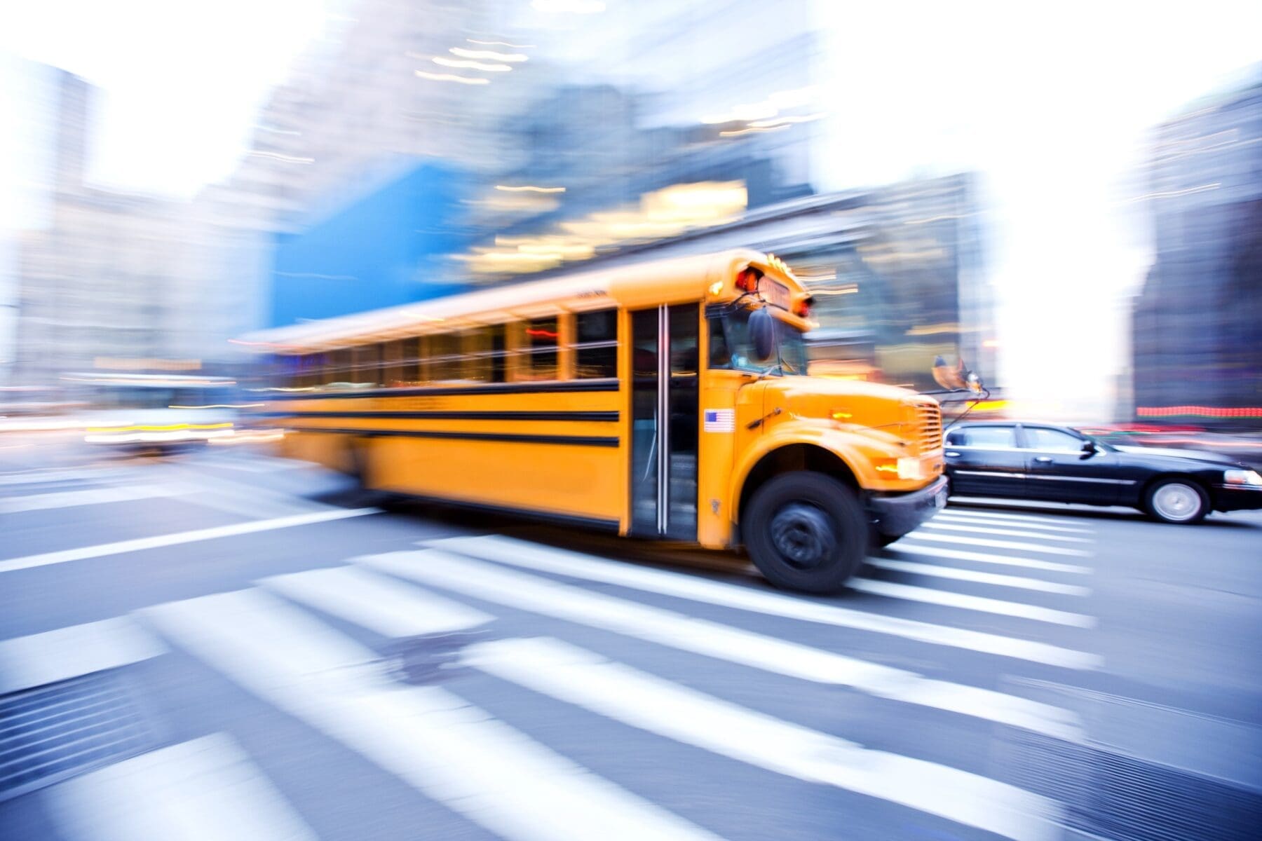 Yellow school bus in motion, driving through a city crosswalk with blurred urban buildings in the background, emphasizing speed and movement.