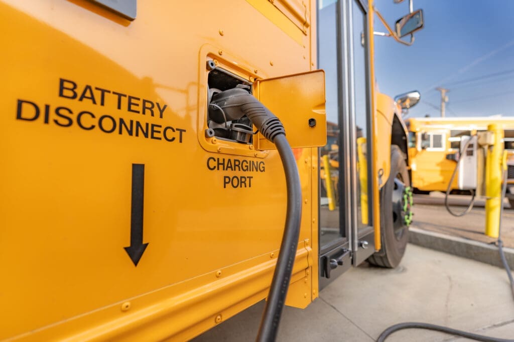 Close-up of an electric school bus being charged at a charging port labeled “Battery Connect” and “Charging Port,” highlighting clean transportation infrastructure.