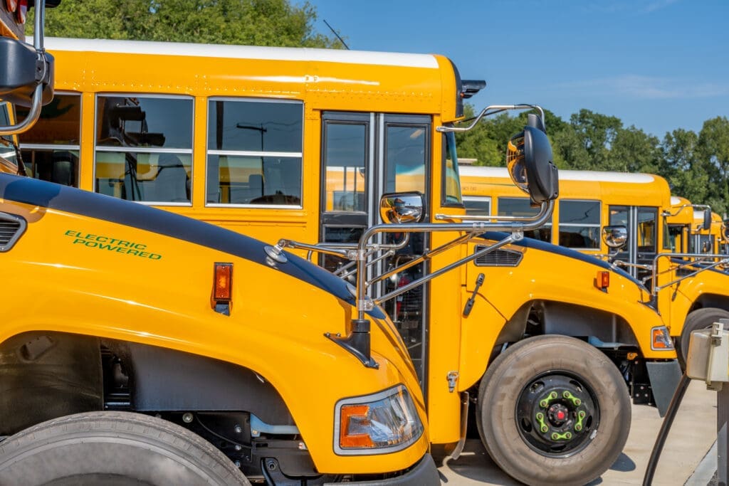 Row of bright yellow electric school buses parked in a lot on a sunny day, with "Electric Powered" text visible on the hood of the nearest bus.