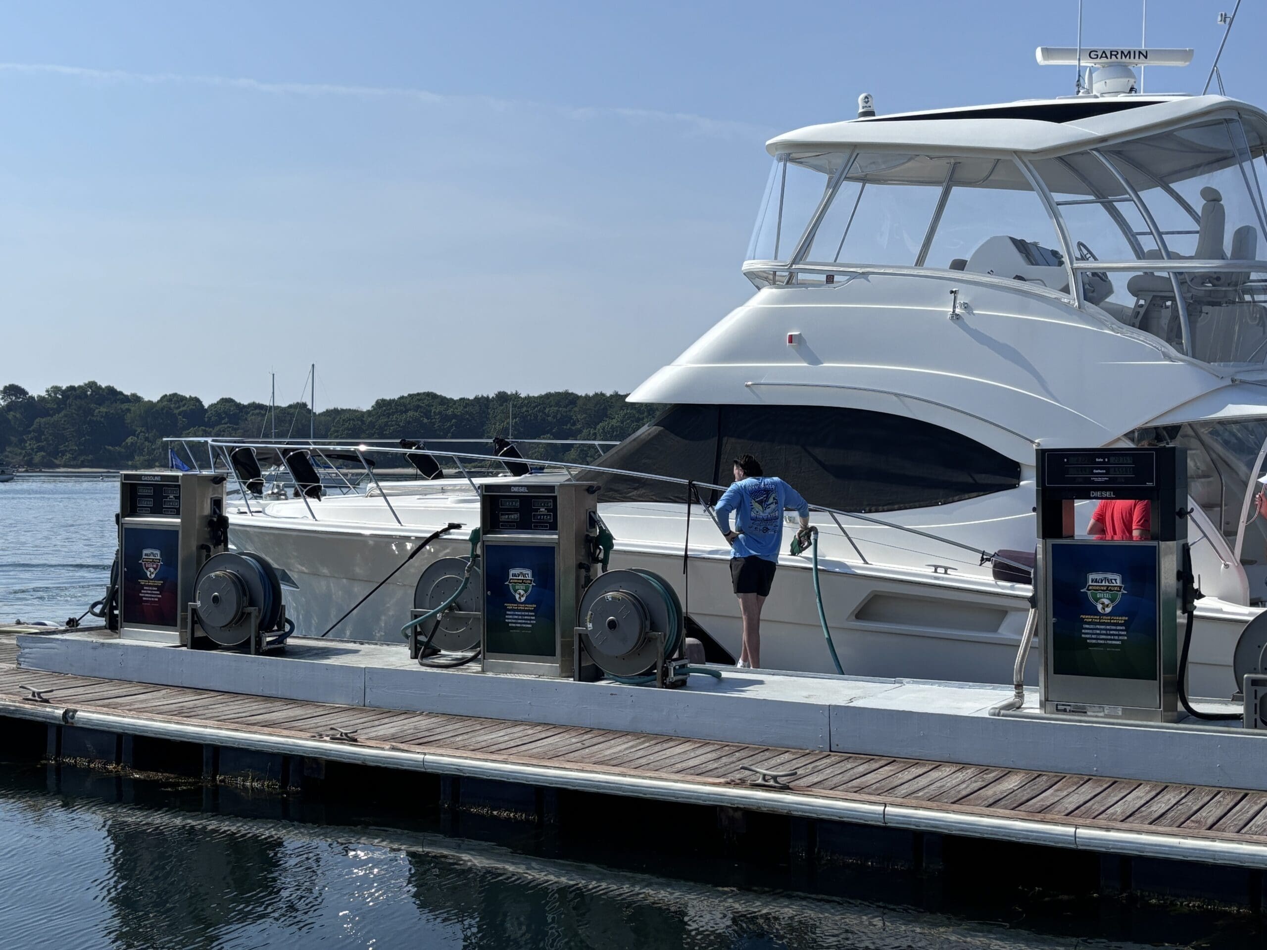 IMG_1998 A white yacht is docked at a marina fueling station, where a person in a blue shirt is refueling the vessel using a hose. The dock is equipped with multiple diesel and gasoline pumps branded with “Harbor Fuels.” Calm waters and a tree-lined shore are visible in the background under a clear blue sky.
